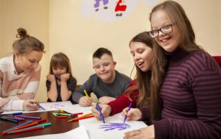 3 young people with disabilities sat at a table drawing with coloured pens, sat with them are two women who are also drawing.