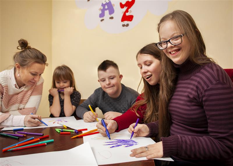 3 young people with disabilities sat at a table drawing with coloured pens, sat with them are two women who are also drawing.